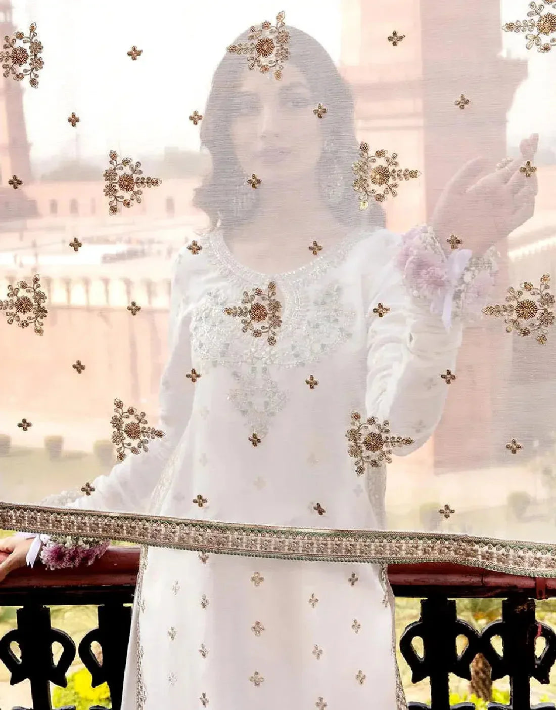 Woman in a white embroidered outfit with gold details, standing in front of a blurred scenic background.