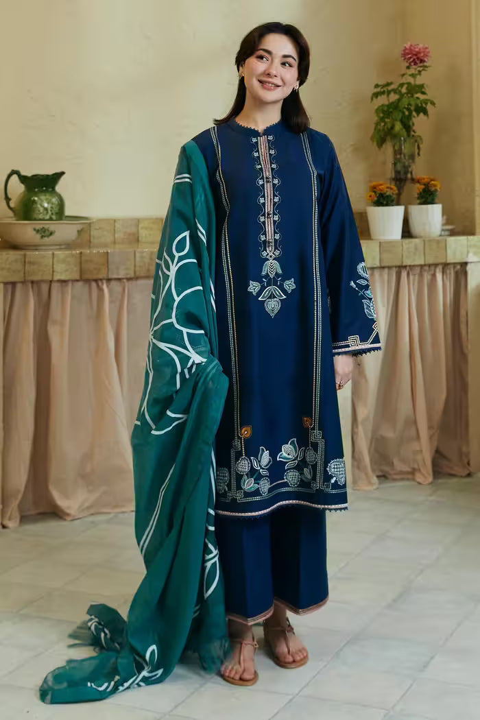 Woman in a blue embroidered outfit with a green dupatta standing indoors.
