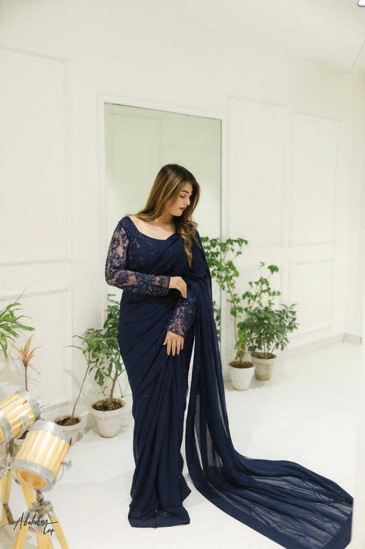Woman in a navy blue saree with lace sleeves standing in a room with plants.