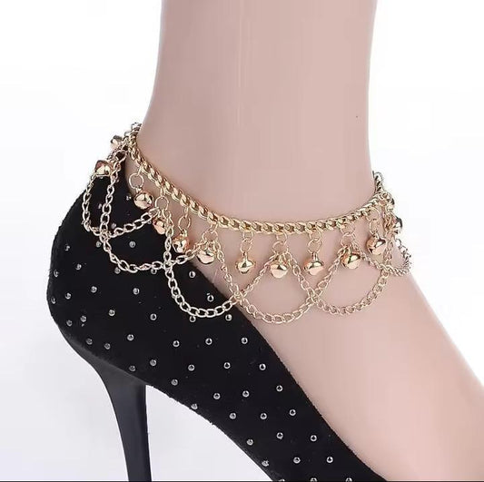 Gold anklet with chains and small bells on a black stiletto shoe against a white background