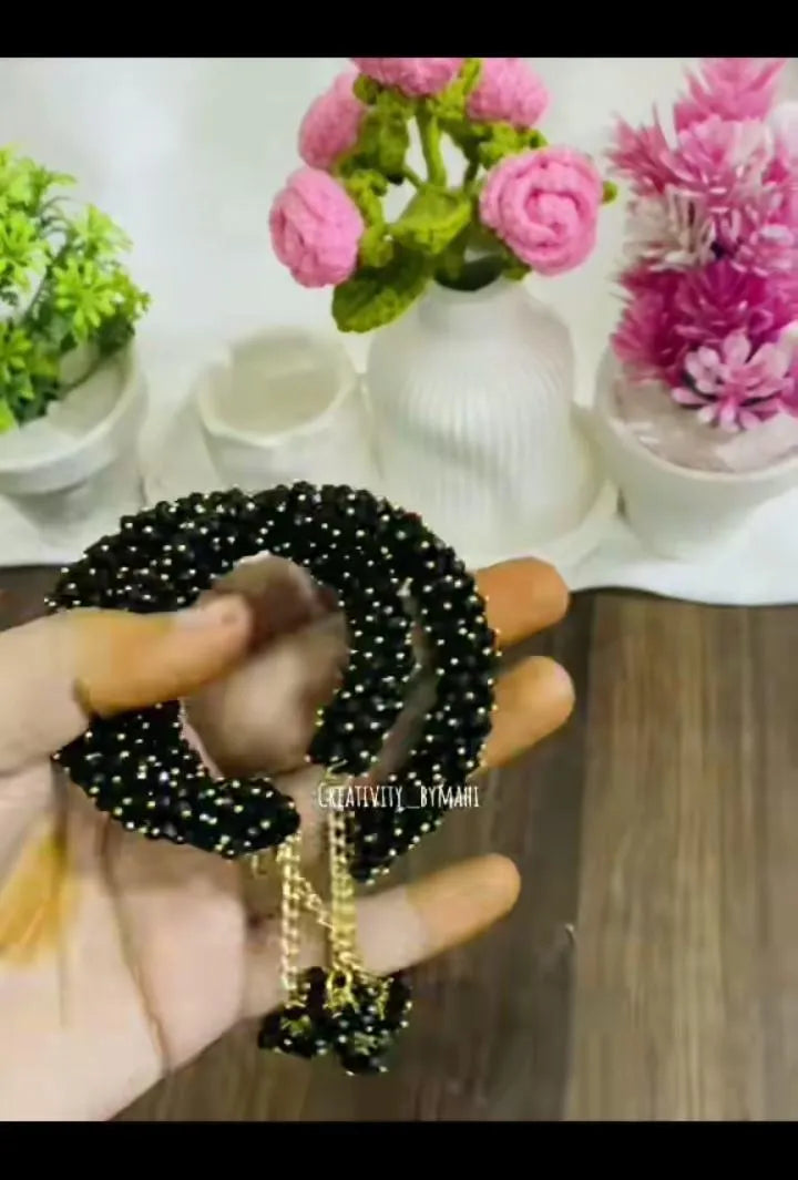 Black hair tie with gold accents held by a hand, with decorative flowers and plants in the background.
