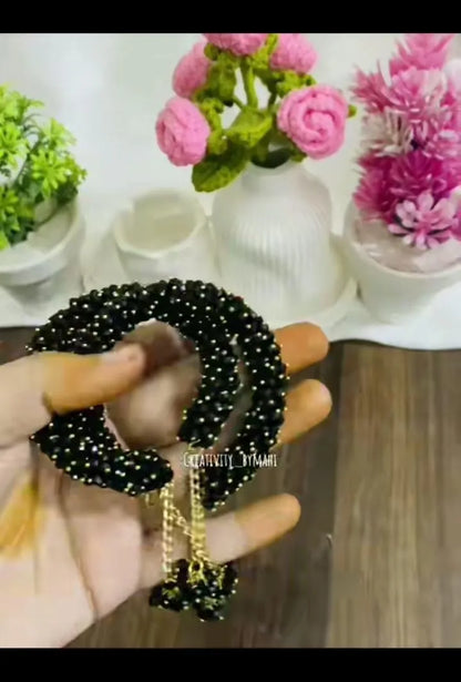 Black hair tie with gold accents held by a hand, with decorative flowers and plants in the background.
