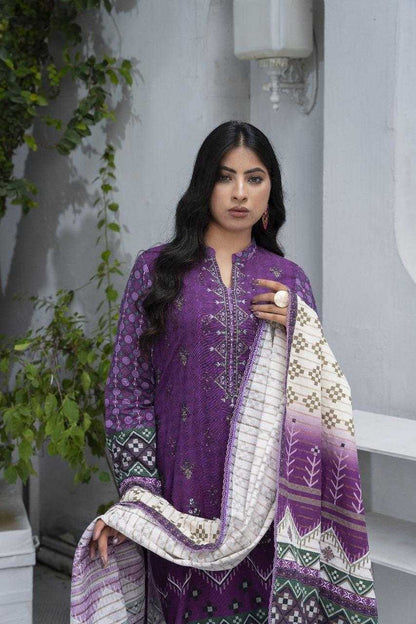 Woman wearing a purple traditional outfit with a white and purple dupatta, standing against a gray wall with greenery.